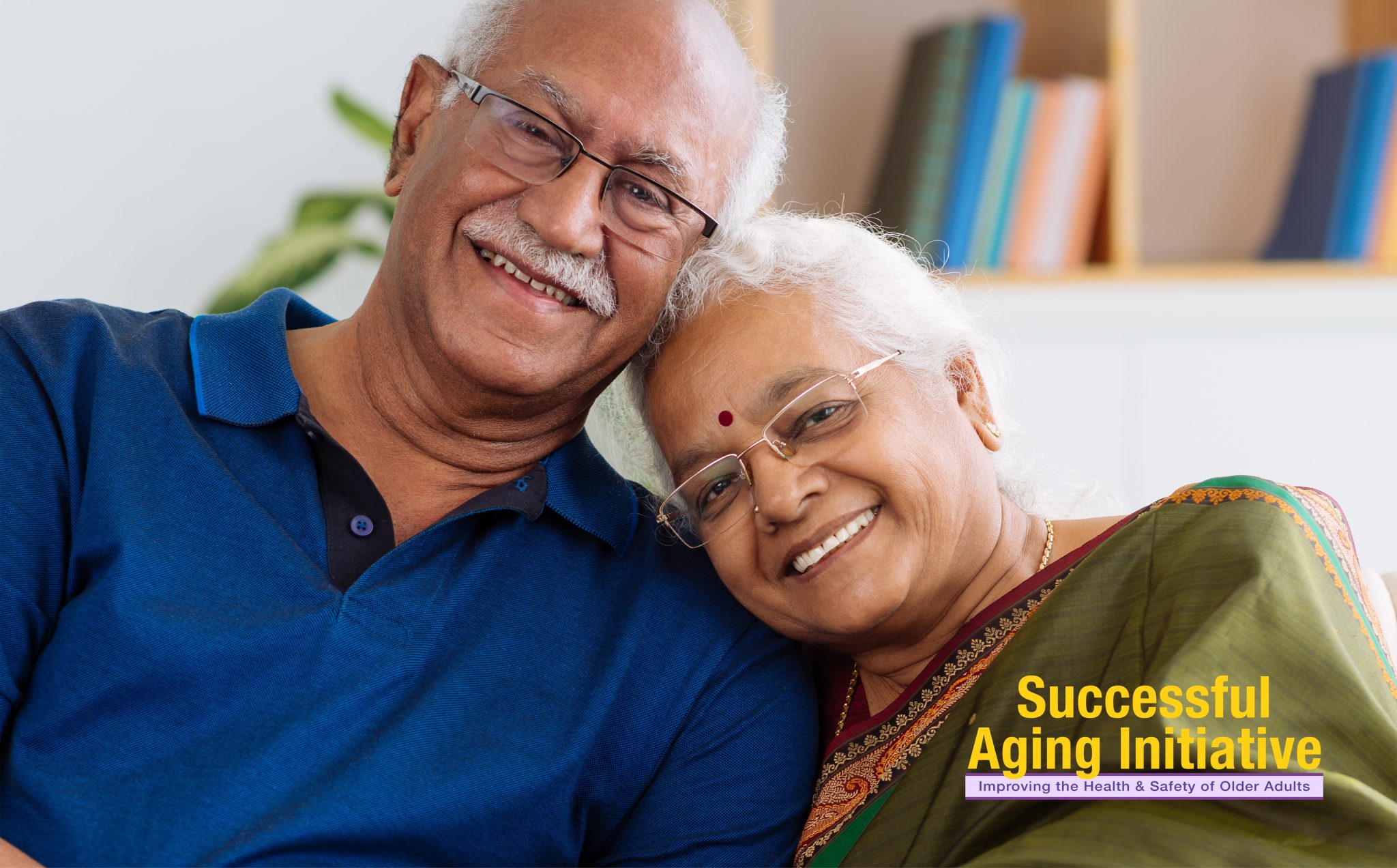 An Indian American older adult couple sits on their sofa, smiling.