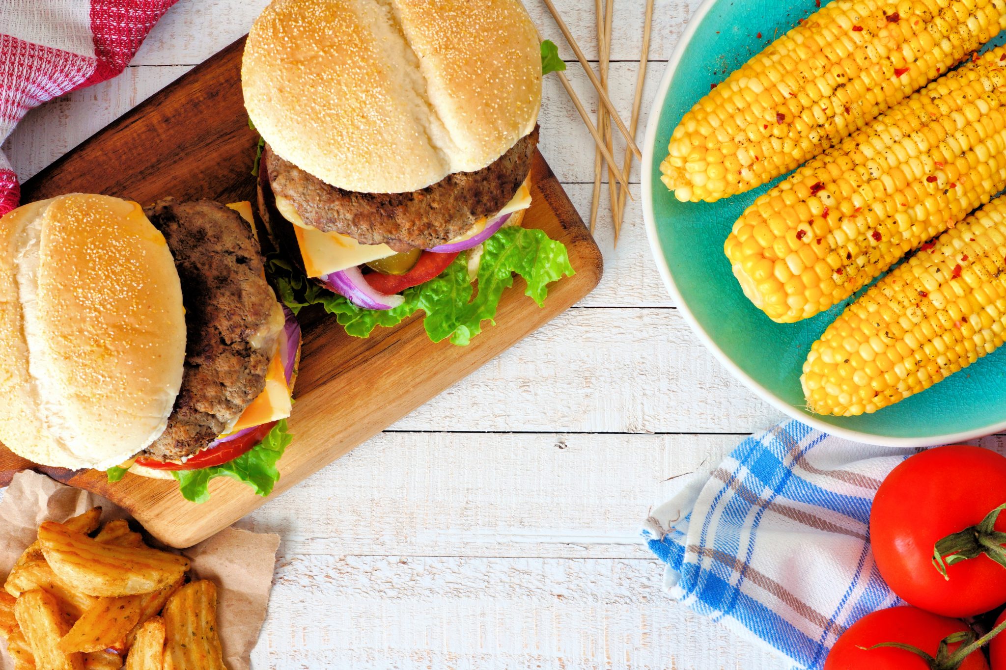 Picnic scene with hamburgers, corn on the cob and potato wedges. Top view over a white wood background.