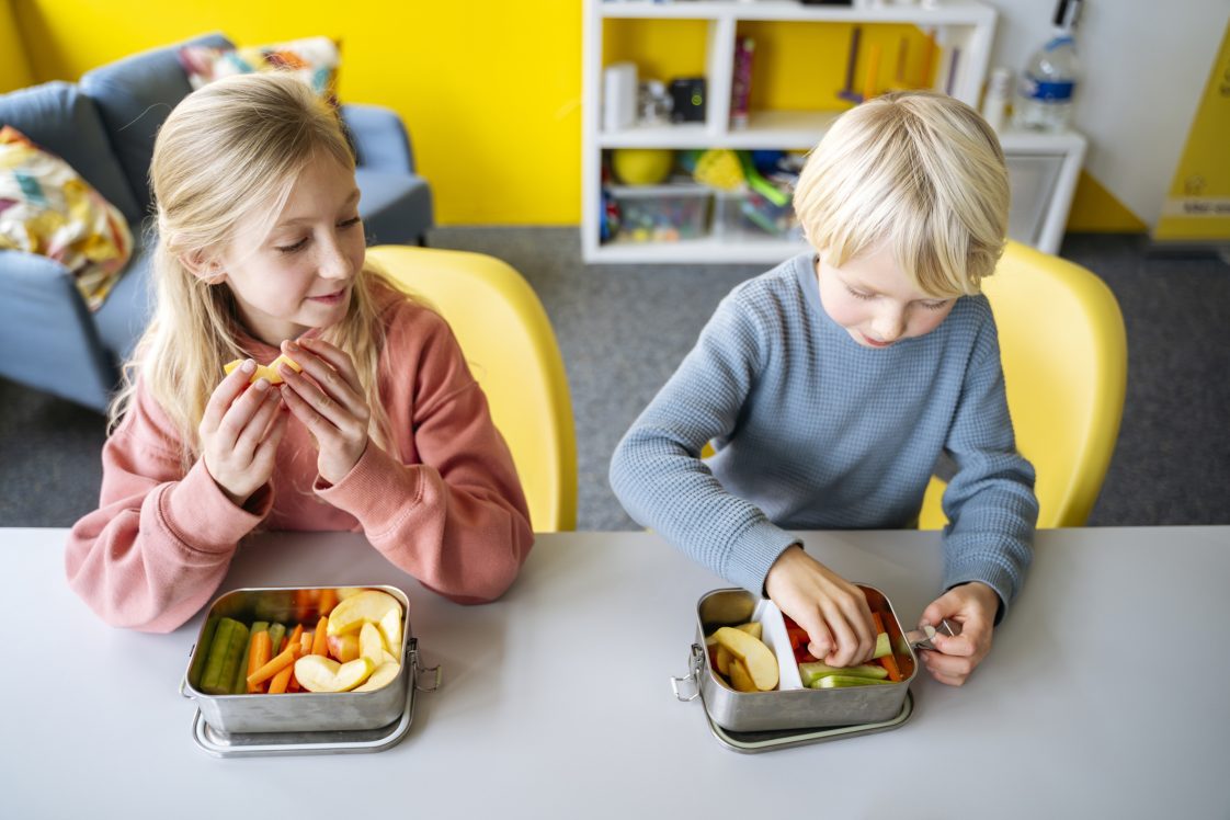 Blond boy and girl eating fruits and vegetables in lunch box at desk