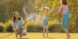 A mother and two daughters playing in the yard, with the mother spraying the girls with a water hose.