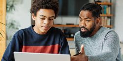 A father helping his teenage son with homework on a computer.