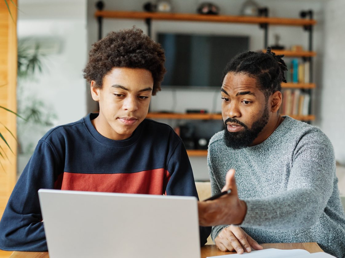 A father helping his teenage son with homework on a computer.