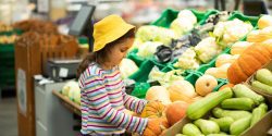 A younger girl in a supermarket/grocery store pickup up a smaller pumpkin.