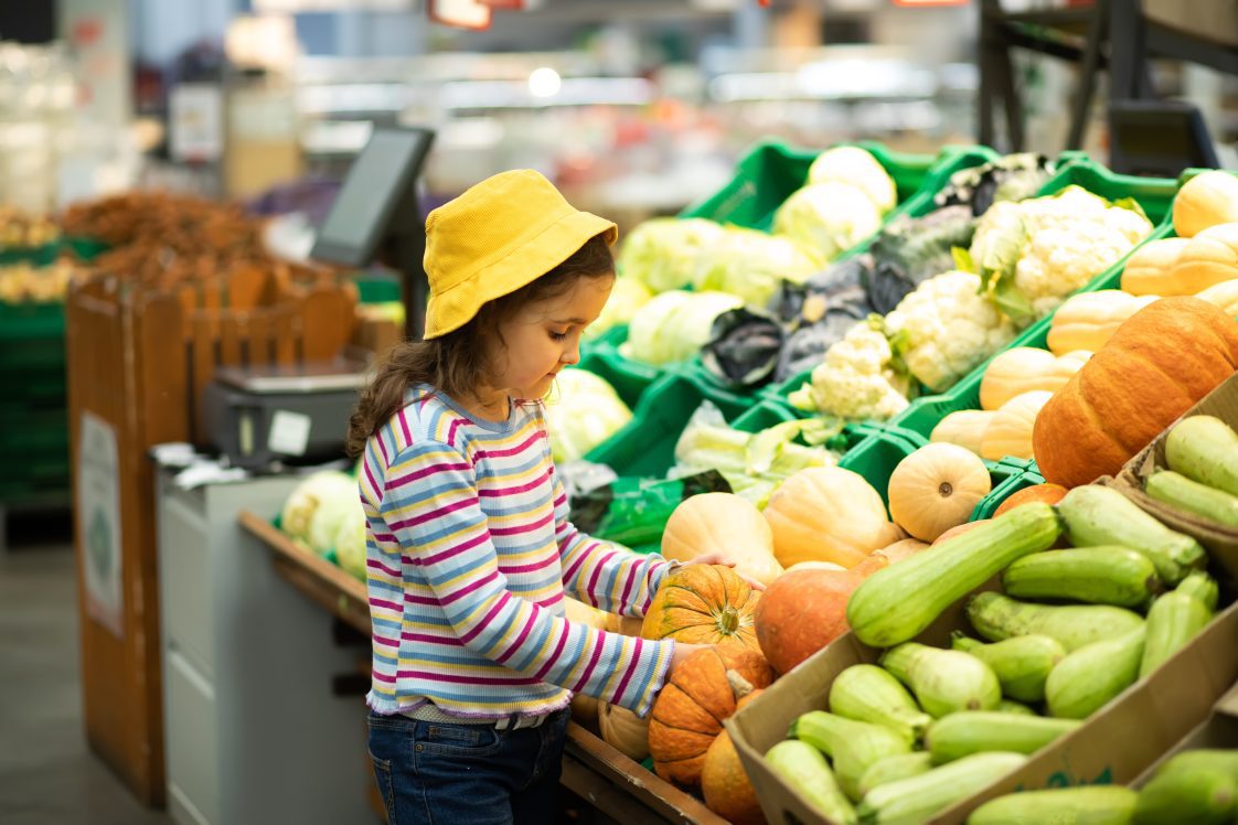 A younger girl in a supermarket/grocery store pickup up a smaller pumpkin.