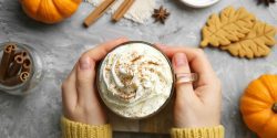 A woman's hands holding a pumpkin spice coffee drink. There are pumpkins, cookies, and spices in the background.