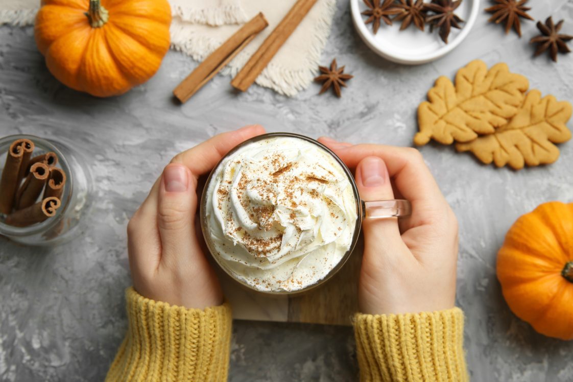 A woman's hands holding a pumpkin spice coffee drink. There are pumpkins, cookies, and spices in the background.