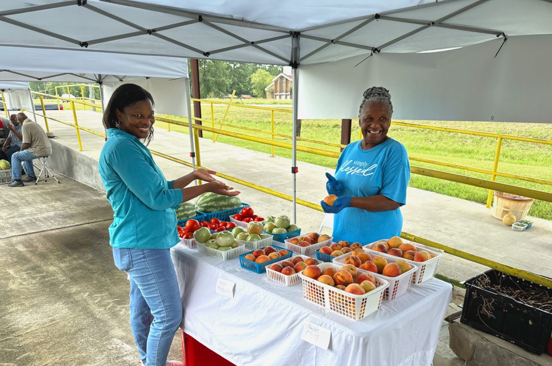 Janice Hall talking with a vendor selling peaches, tomatoes, and watermelon.