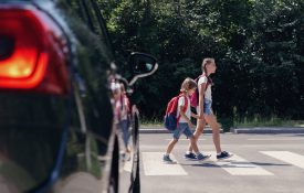 Two kids walking in a crosswalk in front of a car.