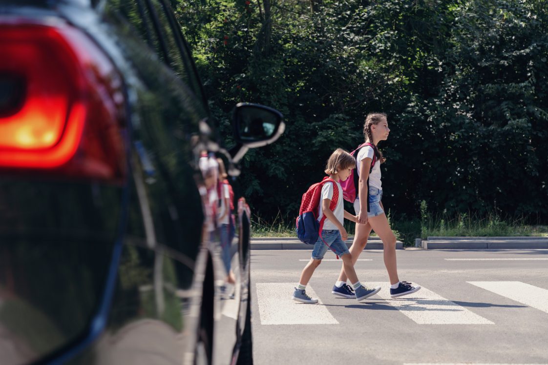 Two kids walking in a crosswalk in front of a car.