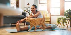 A young Black woman sitting on a yoga mat in her living room and petting an orange cat.