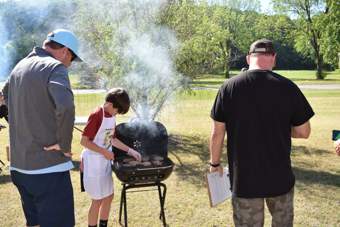 A 4-H member cooking chicken on a grill outdoors.