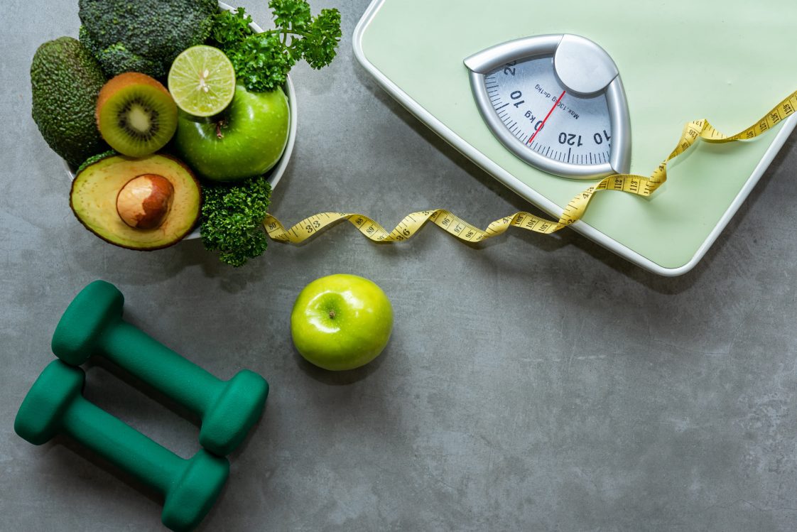 A gray background with a scale, fresh fruits and vegetables, and small weights.