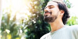 A bearded man is meditating outdoor in the park with face raised up to sky and eyes closed on sunny summer day.