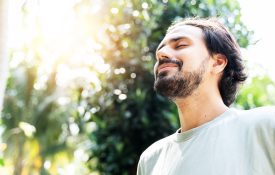 A bearded man is meditating outdoor in the park with face raised up to sky and eyes closed on sunny summer day.