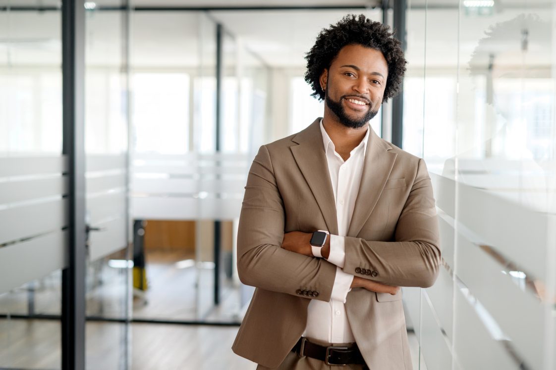 An African-American businessman stands confidently in an office setting with his arms crossed, exuding leadership and professionalism, open office environment on the background